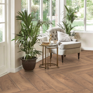 Soggiorno elegante con pavimento Valvia Brown rettangolare effetto legno marrone caldo posa a spina di pesce, poltrona beige, tavolino dorato e piante tropicali con ampia finestra luminosa